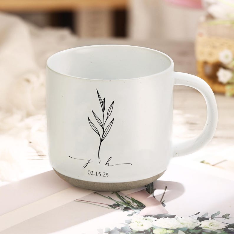 Op de afbeelding: Witte keramische mok met een gespikkelde grijze basis en een gebogen handvat. De mok heeft een zwarte botanische illustratie en de initialen "j + h" met de datum "02.15.25" in een sierlijk schrift. De mok is ontworpen voor dranken.