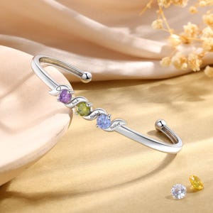 May include: A silver cuff bracelet with three colorful gemstones and engraved names: Nick, Mabel, and Talbot. The bracelet features a curved design with a smooth, polished finish. The gemstones are purple, green, and blue.
