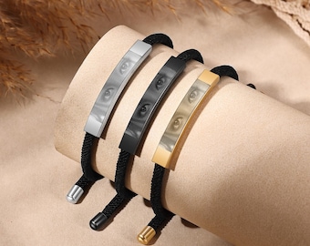 May include: Three bracelets with black rope bands and rectangular metal bars. Each bar features an eye image. The metal bars are silver, black, and gold. The bracelets are displayed on a beige surface.