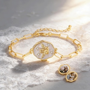 Op de afbeelding: Goudkleurige armband met een rechthoekige schakelketting en een centraal medaillon. Het medaillon heeft een witte emaille achtergrond met een gouden engelontwerp en kleine kristallen. Twee open medaillons met foto's zijn ook zichtbaar.