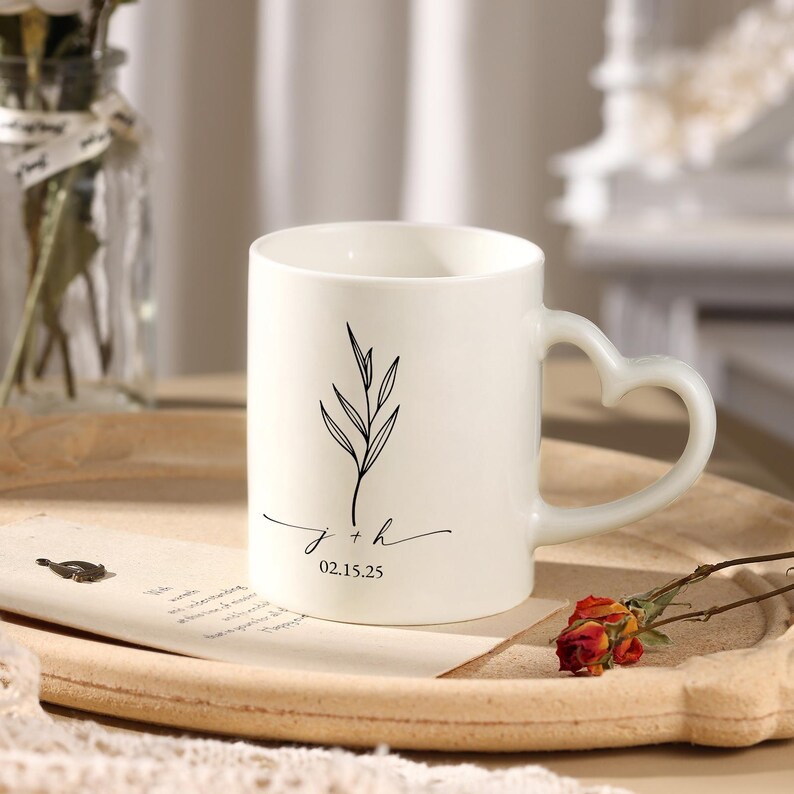 Op de afbeelding: Witte keramische mok met een hartvormig handvat. De mok heeft een zwart botanisch ontwerp en de initialen "j + h" met de datum "02.15.25" in een sierlijk schrift. De mok staat op een houten dienblad.