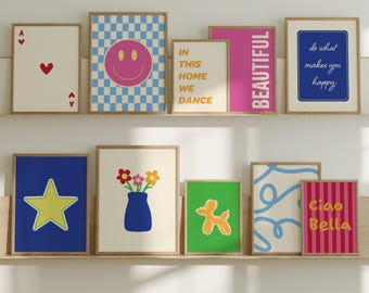 Puede incluir: Una colección de impresiones artísticas enmarcadas exhibidas en dos estantes de madera. Las impresiones presentan varios diseños, incluyendo una carita sonriente, una estrella, flores en un jarrón y textos que dicen "BEAUTIFUL" y "Ciao Bella". La paleta de colores incluye azul, rosa y amarillo.