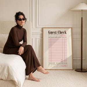 May include: A framed print of a vintage-style guest check with a pink grid and the words "Guest Check" at the top. The print is in a light-colored frame and is displayed on a wall. A woman in a brown turtleneck dress is sitting on a bed.