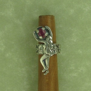 May include: A silver ring with a detailed design of a woman holding a red gemstone. The ring is set on a wooden surface.