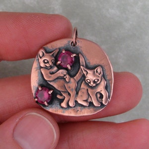 Puede incluir: Colgante de cobre con dos gatitos juguetones con piedras preciosas rosas. El colgante tiene forma ovalada y una superficie texturizada.