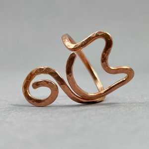 May include: A hammered copper ring with a unique, abstract design. The ring is made of a single piece of wire that is twisted and shaped into a flowing, organic form.