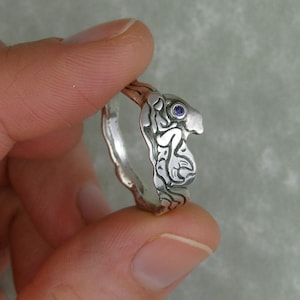 May include: A silver ring with an intricate design and a small purple gemstone. The ring is worn on a finger.