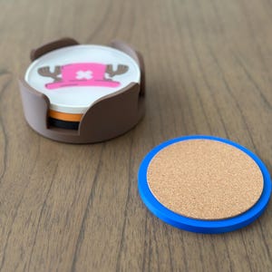 Pode incluir: Conjunto de quatro  apoios de copo marrons e brancos com uma imagem de desenho animado de um chap&eacute;u rosa com uma cruz branca e dois chifres. Os apoios de copo est&atilde;o num suporte castanho. Tamb&eacute;m &eacute; mostrado um apoio de copo separado de corti&ccedil;a e azul.