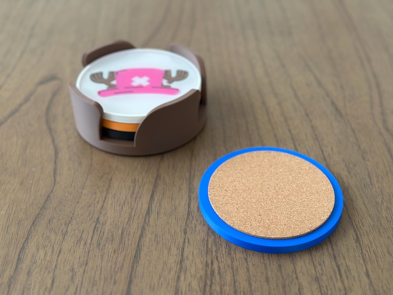 Pode incluir: Conjunto de quatro  apoios de copo marrons e brancos com uma imagem de desenho animado de um chap&eacute;u rosa com uma cruz branca e dois chifres. Os apoios de copo est&atilde;o num suporte castanho. Tamb&eacute;m &eacute; mostrado um apoio de copo separado de corti&ccedil;a e azul.