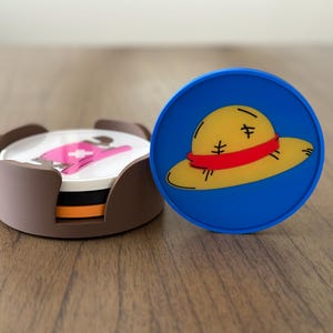 May include: Set of four brown coasters with a pink and white design. A blue coaster with a yellow straw hat with a red band.