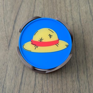 Pode incluir: Um coaster redondo azul e marrom com uma imagem de desenho animado de um chap&eacute;u de palha amarelo com uma faixa vermelha.