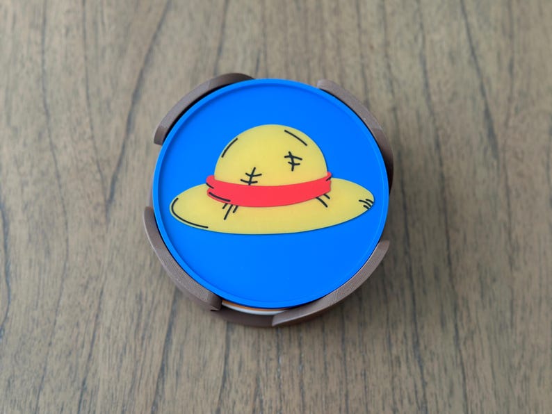 Pode incluir: Um coaster redondo azul e marrom com uma imagem de desenho animado de um chap&eacute;u de palha amarelo com uma faixa vermelha.