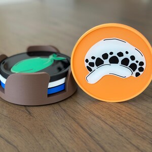 Pode incluir: Dois porta-copos redondos de silicone. Um porta-copos &eacute; laranja com uma imagem de desenho animado em preto e branco de uma caveira com um &uacute;nico olho. O outro porta-copos &eacute; marrom com um design de peixe verde e faz parte de um conjunto de quatro porta-copos.