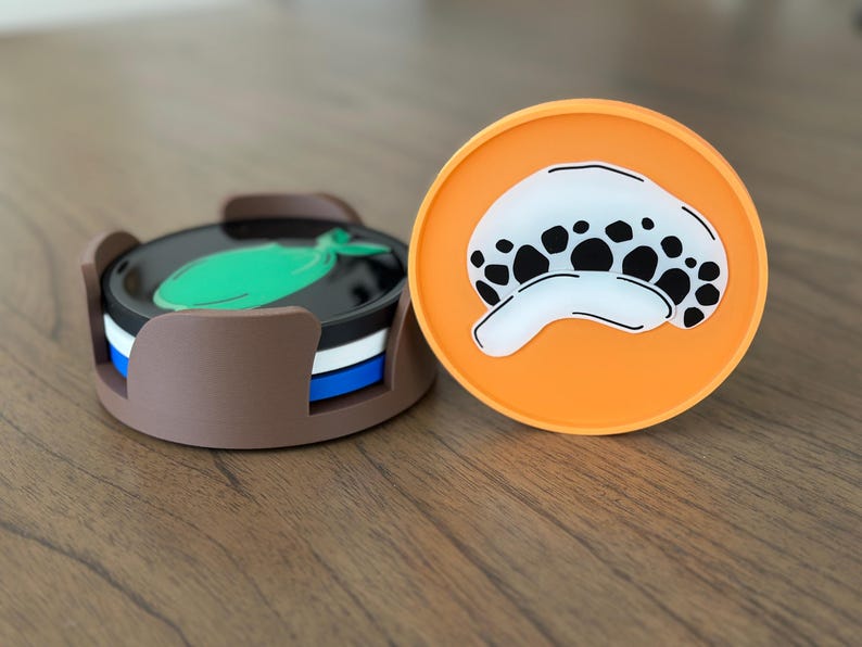Pode incluir: Dois porta-copos redondos de silicone. Um porta-copos &eacute; laranja com uma imagem de desenho animado em preto e branco de uma caveira com um &uacute;nico olho. O outro porta-copos &eacute; marrom com um design de peixe verde e faz parte de um conjunto de quatro porta-copos.