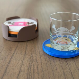 Pode incluir: Dois  coasters com um design de caveira e ossos cruzados. Um coaster &eacute; marrom e tem uma pilha de tr&ecirc;s coasters. O outro coaster &eacute; azul e tem um design amarelo. Um copo com um design de caveira e ossos cruzados est&aacute; sobre o coaster azul.