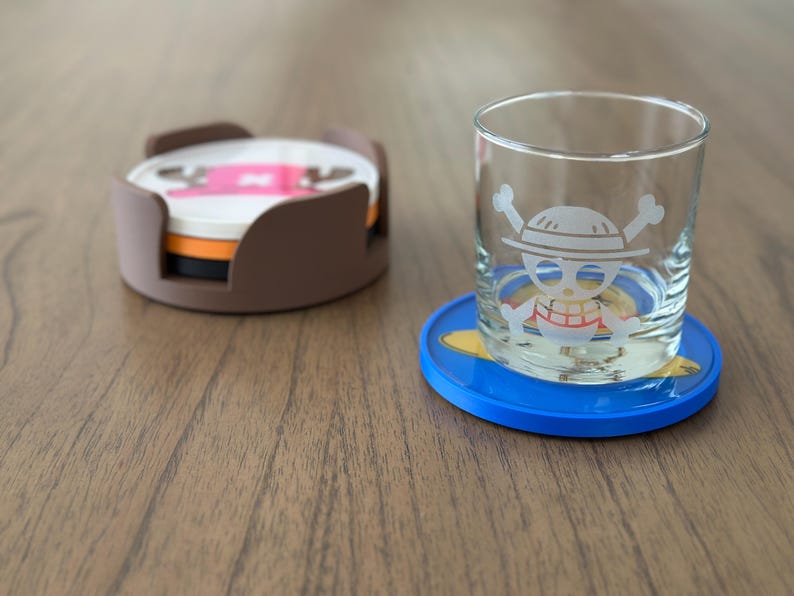 Pode incluir: Dois  coasters com um design de caveira e ossos cruzados. Um coaster &eacute; marrom e tem uma pilha de tr&ecirc;s coasters. O outro coaster &eacute; azul e tem um design amarelo. Um copo com um design de caveira e ossos cruzados est&aacute; sobre o coaster azul.