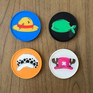 Pode incluir: Quatro  tapetes redondos com diferentes personagens de desenhos animados. Os tapetes s&atilde;o azuis, pretos, laranja e brancos. Os personagens s&atilde;o um chap&eacute;u de palha, uma bandana verde, uma caveira com manchas pretas e um chap&eacute;u rosa com chifres.