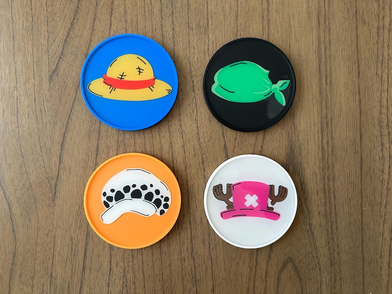 Pode incluir: Quatro  tapetes redondos com diferentes personagens de desenhos animados. Os tapetes s&atilde;o azuis, pretos, laranja e brancos. Os personagens s&atilde;o um chap&eacute;u de palha, uma bandana verde, uma caveira com manchas pretas e um chap&eacute;u rosa com chifres.