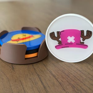 Pode incluir: Dois  copos de apoio com personagens do anime One Piece. Um copo de apoio &eacute; um suporte castanho com quatro copos de apoio azuis, laranja e amarelos no seu interior. Os copos de apoio representam um chap&eacute;u de palha com uma faixa amarela. O outro copo de apoio &eacute; branco com um chap&eacute;u rosa com uma cruz branca e chifres castanhos.