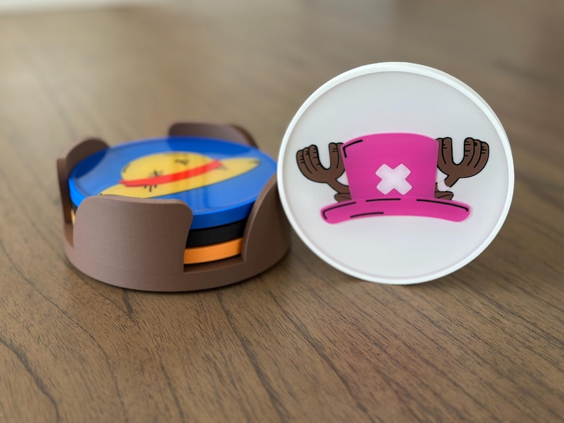 Pode incluir: Dois  copos de apoio com personagens do anime One Piece. Um copo de apoio &eacute; um suporte castanho com quatro copos de apoio azuis, laranja e amarelos no seu interior. Os copos de apoio representam um chap&eacute;u de palha com uma faixa amarela. O outro copo de apoio &eacute; branco com um chap&eacute;u rosa com uma cruz branca e chifres castanhos.