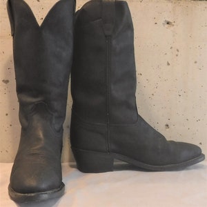 Könnte beinhalten: Ein Paar schwarze Cowboy-Stiefel aus Leder. Die Stiefel haben spitze Zehen, abgeschrägte Absätze und hohe Schäfte. Sie sind im klassischen Western-Stil gehalten und scheinen abgenutzt zu sein. Jeder Stiefel ist etwa 30 cm hoch.