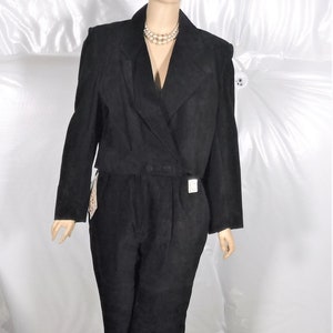 Puede incluir: Mono de ante negro con una parte superior estilo blazer de doble botonadura y pantalones a juego. La blazer tiene solapas con muescas y dos botones. Los pantalones están plisados en la cintura. El maniquí lleva un collar de perlas.