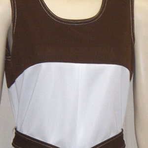 Op de afbeelding: Een mouwloze jurk met een bruin lijfje en een witte rok, met een bijpassende bruine riem die in de taille is vastgeknoopt. De jurk heeft witte stikseldetails en een ronde halslijn. De algehele stijl doet denken aan vintage mode.