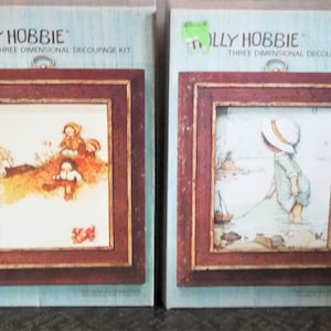 Może przedstawiać: Dwa zestawy do decoupage'u trójwymiarowego Holly Hobbie w stylu vintage. Każdy zestaw zawiera oprawioną ilustrację z brązową drewnianą ramą. Jeden przedstawia bawiące się dzieci, drugi dziecko łowiące ryby. Zestawy są na jasnoniebieskim tle.
