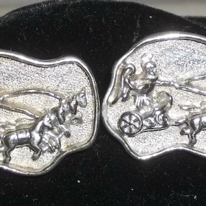 May include: A pair of silver-toned cufflinks. Each cufflink features a detailed relief of a chariot pulled by horses, with a figure in the chariot. The background has a textured appearance. The cufflinks are set against a dark background.