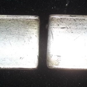Op de afbeelding: Twee zilverkleurige, rechthoekige manchetknopen. Elke manchetknop heeft een licht afgeronde rand en een geborstelde metalen afwerking. De manchetknopen worden gepresenteerd tegen een donkere achtergrond, wat hun metalen glans benadrukt.