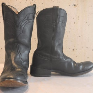 Könnte beinhalten: Ein Paar schwarze Leder-Cowboystiefel. Die Stiefel haben ein klassisches Design mit dekorativen Nähten am Schaft und einer spitzen Zehenpartie. Die Sohlen weisen Gebrauchsspuren auf. Klassischer Western-Stil.