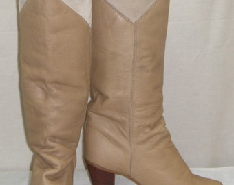 May include: A pair of tan leather cowboy boots with a light beige suede upper section. The boots have a pointed toe, a wooden heel, and metal accents on the toe. The boots are knee-high.