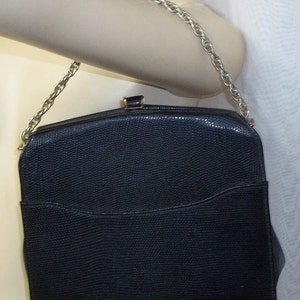 Puede incluir: Un bolso de mano azul marino oscuro con una correa de cadena plateada. El bolso tiene un patrón texturizado, similar a la piel de reptil, y una forma rectangular con una parte superior curvada. Un pequeño bolsillo es visible en la parte delantera.