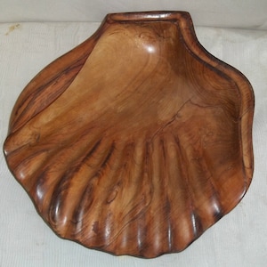 Puede incluir: Un cuenco de madera con forma de concha. Elaborado en madera marrón claro con vetas más oscuras, presenta bordes festoneados y un interior liso y pulido. Este cuenco decorativo imita la forma de una gran concha marina.