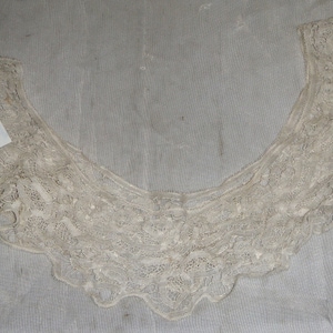 Puede incluir: Cuello de encaje color crema con borde festoneado. El cuello tiene un diseño calado delicado y una etiqueta con el texto "Belles, Cotton, needle lace, c. 1890".
