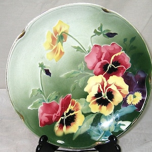Peut inclure: Une assiette décorative avec un fond vert et un motif floral représentant des pensées aux tons de jaune, rouge et violet. L'assiette a un bord festonné avec une bordure dorée et est présentée sur un support noir.