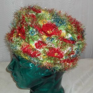 May include: A colorful, handmade hat with a floral pattern. The hat features a base fabric with red, yellow, blue, and green floral designs, and is trimmed with a fluffy, multi-colored yarn. The hat is displayed on a green mannequin head.