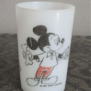May include: White glass tumbler featuring a black and white illustration of Mickey Mouse holding a bone. Mickey Mouse is wearing a white shirt and red pants. The tumbler has the text "Walt Disney Productions" at the bottom.