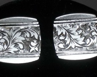 Könnte beinhalten: Ein Paar silberne Manschettenknöpfe mit einem gravierten floralen Design. Die rechteckigen Manschettenknöpfe haben abgerundete Kanten und ein detailliertes Muster aus Blättern und Ranken. Das Design ist in das Metall geätzt und erzeugt ein strukturiertes Aussehen.