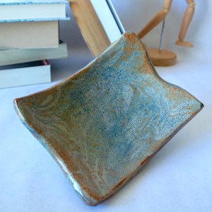 May include: A handmade ceramic dish with a square shape and gently curved edges. The dish features a mottled blue and brown glaze, creating a textured appearance. The dish is approximately 6 inches wide.