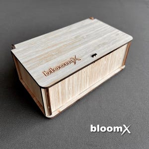 May include: A wooden box with a light brown finish and the word "bloomx" engraved on the top. The box has a hinged lid and a small metal latch.