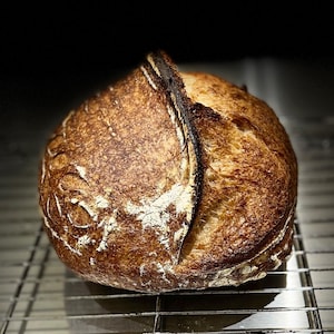 May include: A freshly baked loaf of sourdough bread with a dark brown crust and a light brown interior. The bread is sitting on a wire rack.