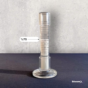 May include: A clear glass graduated cylinder with a white liquid inside. The liquid level is at the 75% mark. The cylinder is labeled "bloomX".