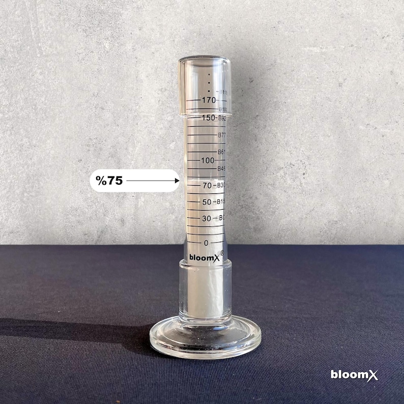 May include: A clear glass graduated cylinder with a white liquid inside. The liquid level is at the 75% mark. The cylinder is labeled "bloomX".