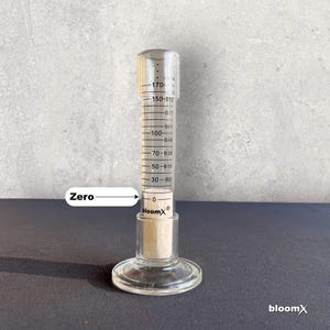 May include: A clear glass graduated cylinder with a white plastic stopper. The cylinder has markings in milliliters and is labeled "bloomX&reg;". The cylinder is filled with a clear liquid up to the 30 ml mark. The word "Zero" is written on the left side of the image with an arrow pointing to the 0 ml mark on the cylinder.