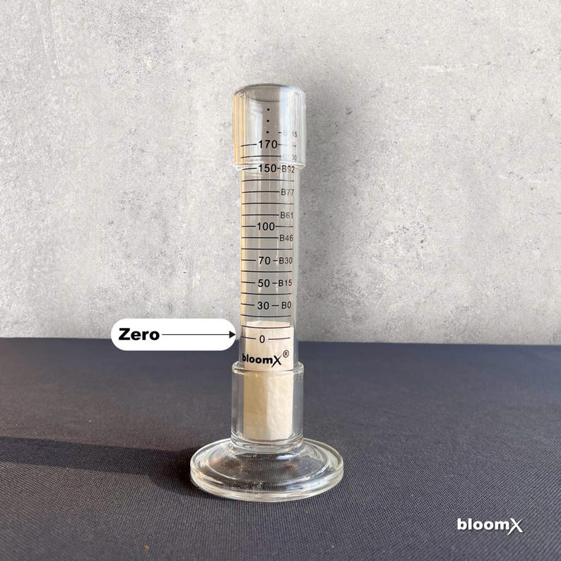 May include: A clear glass graduated cylinder with a white plastic stopper. The cylinder has markings in milliliters and is labeled "bloomX&reg;". The cylinder is filled with a clear liquid up to the 30 ml mark. The word "Zero" is written on the left side of the image with an arrow pointing to the 0 ml mark on the cylinder.