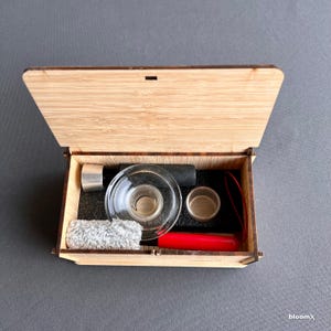 May include: A wooden box with a hinged lid, open to reveal a glass bowl, a metal pipe, a small red tool, a gray cleaning cloth, and two small metal containers. The box is lined with black foam.