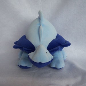 May include: A blue and purple plush toy shaped like a dinosaur. The dinosaur is lying on its belly with its head and tail raised.