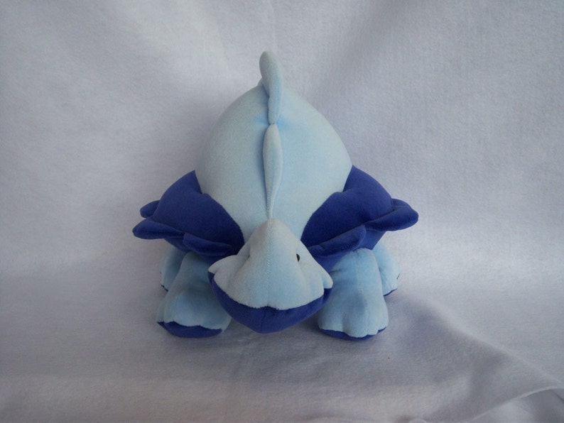 May include: A blue and purple plush toy shaped like a dinosaur. The dinosaur is lying on its belly with its head and tail raised.