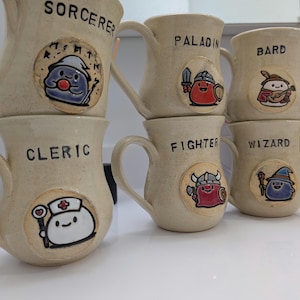 Op de afbeelding: Een stapel beige keramische mokken, elk met een uniek stripfiguur en klassenaam: Sorcerer, Paladin, Bard, Cleric, Fighter en Wizard. De mokken hebben gebogen handvatten en zijn versierd met kleurrijke illustraties.